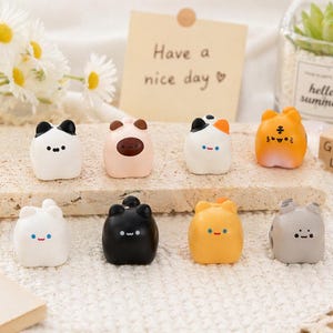 May include: Eight small, colourful cat-shaped figurines. The figurines are white, black, orange, and grey, with painted faces. A note saying "Have a nice day" is in the background.