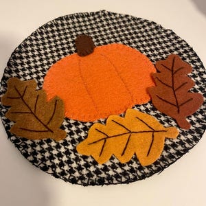 Op de afbeelding: Een ronde stoffen onderzetter met een zwart-wit pied-de-poulepatroon. De onderzetter is versierd met een centrale oranje viltpompoen met een bruine steel, geflankeerd door drie vilten bladeren in de kleuren bruin, oranje en geel.