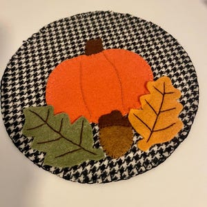Könnte beinhalten: Ein runder Untersetzer aus Stoff mit schwarz-weißem Hahnentrittmuster. Die Mitte ist mit einem leuchtend orangefarbenen Filzkürbis, einer kleinen braunen Filzeichel, einem grünen Filz-Eichenblatt und einem gelb-orangen Filzblatt mit Stickereien verziert.