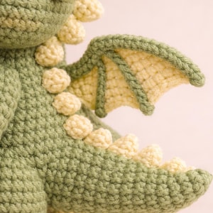 Puede incluir: Drag&oacute;n verde tejido a ganchillo con detalles amarillos. El peluche hecho a mano presenta un cuerpo texturizado, peque&ntilde;as esferas amarillas a lo largo de la espalda y un ala detallada. El ala es de color amarillo claro con un borde verde.
