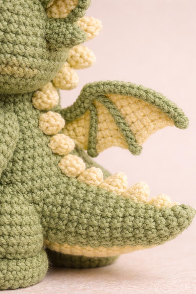 Puede incluir: Drag&oacute;n verde tejido a ganchillo con detalles amarillos. El peluche hecho a mano presenta un cuerpo texturizado, peque&ntilde;as esferas amarillas a lo largo de la espalda y un ala detallada. El ala es de color amarillo claro con un borde verde.