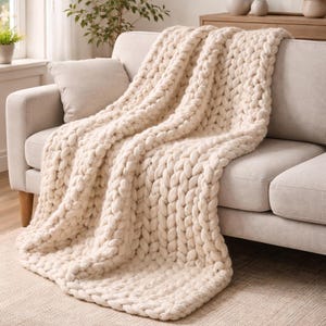 Puede incluir: Una manta de punto grueso de color crema sobre un sofá beige claro. Las puntadas gruesas y de gran tamaño de la manta crean una apariencia texturizada y acogedora. El sofá está sobre una alfombra de tonos neutros, con una ventana y plantas en el fondo. Es un artículo de decoración.
