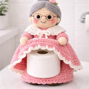 May include: A crocheted toilet roll holder in the shape of a grandmother. The figure has grey hair, glasses, and a pink dress with white lace trim. A roll of toilet paper is held in the dress.