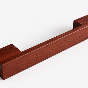 May include: A rectangular, solid wood cabinet pull with a rich, reddish-brown finish. The hardware features a handle with two square end pieces, creating a modern, minimalist design. The pull is designed for easy grip and installation.