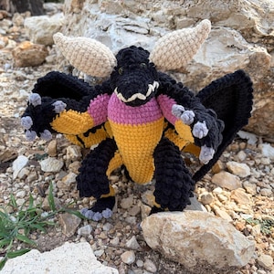 May include: A crocheted dragon plush toy featuring a black body, yellow chest, and pink accents. The creature has large beige ears, dark eyes, a white-toothed grin, and grey claws on its hands and feet. It has wide, dark, textured wings.