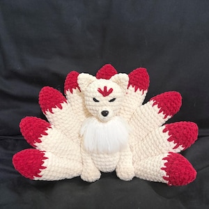 Puede incluir: Un peluche de zorro hecho a mano con nueve colas. El zorro es principalmente de color crema con detalles rojos en las puntas de las colas y las orejas. Tiene un pecho blanco y esponjoso y una marca roja en la frente.