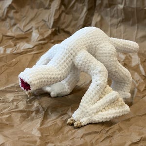 May include: A white crocheted creature with multiple limbs and a textured surface. The creature has a red interior in its mouth and tan-colored feet. The handmade plush toy is displayed on a brown paper background.