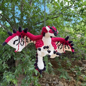 May include: A handmade crochet dragon in red, white, and black. The dragon has large wings with a unique pattern, black spikes, and is suspended against a backdrop of green foliage. The dragon is a whimsical, handcrafted item.