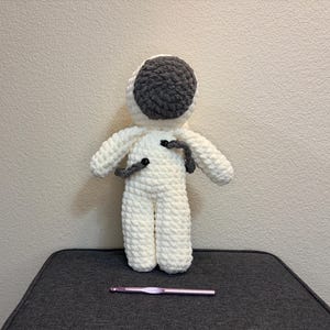 May include: A handmade, white crocheted astronaut doll with a gray helmet. The doll has gray arm details and black button accents. A pink crochet hook rests in front of the doll on a dark gray surface.