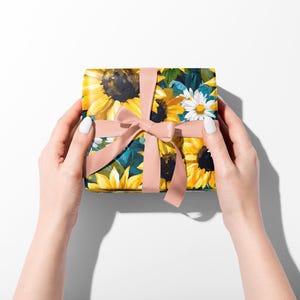 May include: A square gift box wrapped in floral paper featuring sunflowers and daisies, tied with a light pink ribbon. The box is held by two hands with white nail polish against a white background.