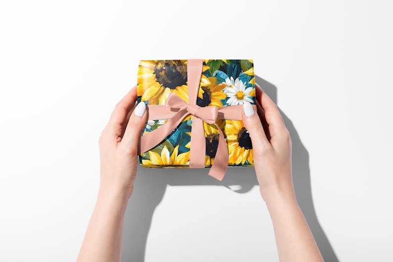 May include: A square gift box wrapped in floral paper featuring sunflowers and daisies, tied with a light pink ribbon. The box is held by two hands with white nail polish against a white background.