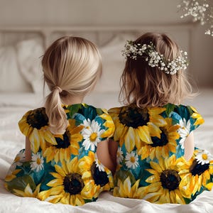 May include: Two young children wearing matching dresses with a sunflower and daisy pattern. The dresses feature yellow sunflowers, white daisies, and teal green leaves. One child has a ponytail, and the other wears a flower crown.