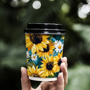 May include: A disposable coffee cup with a black lid, featuring a vibrant floral design of yellow sunflowers and white daisies against a teal background. The cup is held in a person's hand.