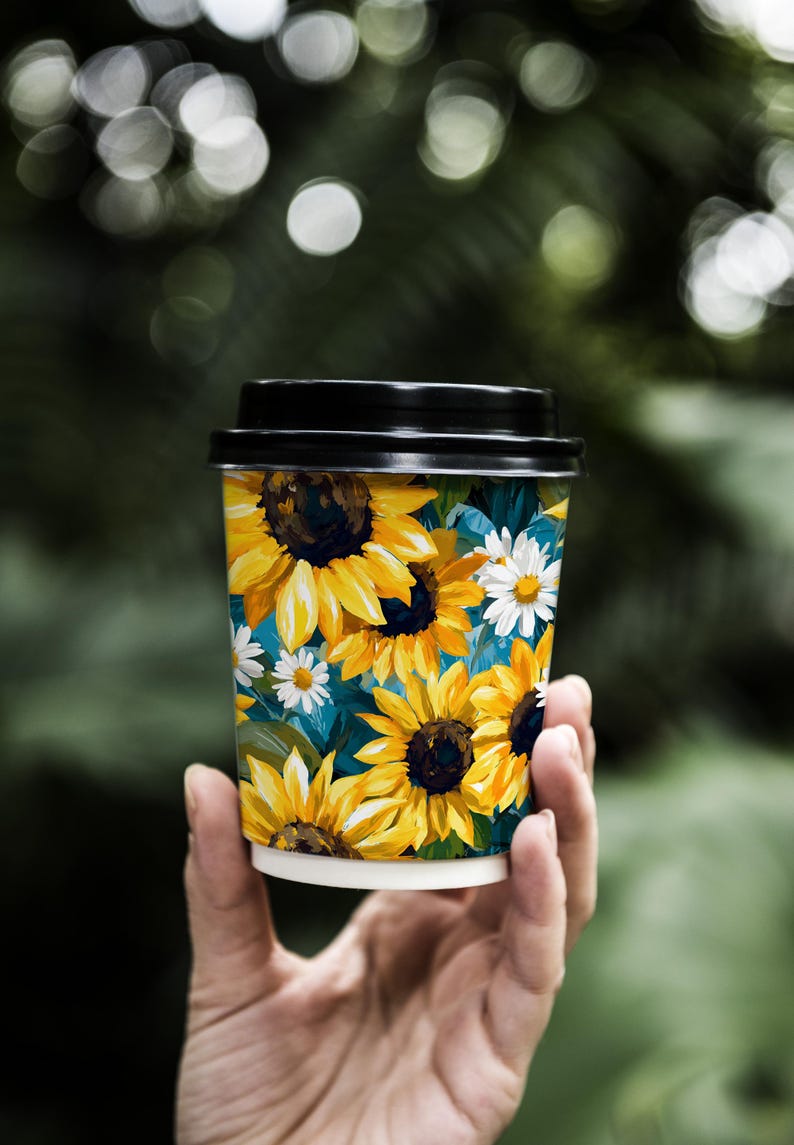 May include: A disposable coffee cup with a black lid, featuring a vibrant floral design of yellow sunflowers and white daisies against a teal background. The cup is held in a person's hand.