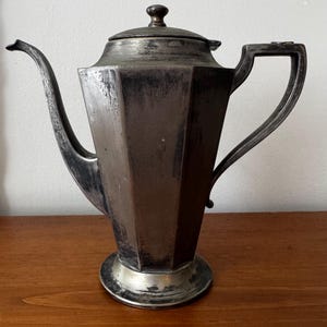 May include: A silver-toned metal coffee pot with a hexagonal body, a long curved spout, and a matching lid with a small knob handle. The vessel features a wide base and a sturdy, angular handle, showing signs of natural oxidation and patina.