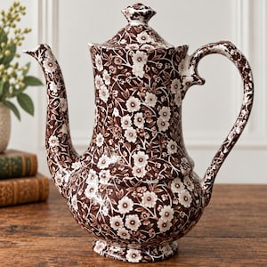 May include: A ceramic teapot featuring a dark brown background with a repeating white floral pattern. The teapot has a curved handle, a long spout, and a matching lid with a small knob on top. The entire surface is covered in the floral design.