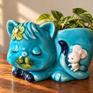 May include: A glossy turquoise ceramic planter shaped like a sleeping cat with a yellow flower on its head and a small white mouse figure attached to its side. The cat has closed eyes, painted whiskers, and dark blue paws, with a green plant inside.