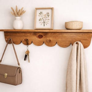 May include: A wooden wall shelf with a scalloped edge, holding a framed botanical print, a woven basket, and a white vase with dried flowers. A taupe purse, keys, and a beige blanket hang below.