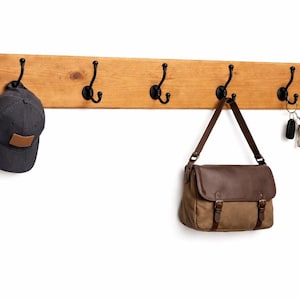 May include: Wooden coat rack with five black metal hooks. A dark gray baseball cap, a brown and tan messenger bag, and a set of keys hang from the hooks. The rack is made of light brown wood.