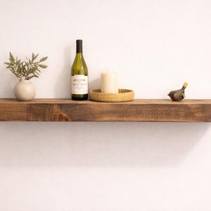 May include: A rustic, dark-stained wooden floating shelf with decorative items. It features a small white vase with greenery, a wine bottle, a candle in a woven holder, and a bird figurine. The shelf is mounted on a white wall.