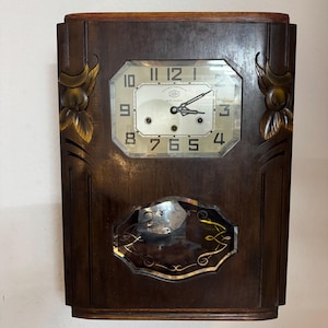 May include: A dark brown wooden wall clock with an octagonal clock face and a rectangular body. The clock face has black numerals and hands. Decorative gold-colored accents are on the sides. The clock has a mirrored lower section.