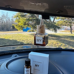 May include: A car air freshener with a wooden top and a glass bottle, suspended by a string. The label reads "MY". A small bottle and a white box with "MY Sandal Amber" are also present.