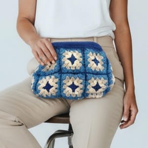 Op de afbeelding: Een blauw en crèmekleurige gehaakte clutch met een bloemenpatroon. De clutch wordt vastgehouden door een persoon die een witte top en een beige broek draagt. De tas heeft een rechthoekige vorm en een donkerblauwe bovenkant.
