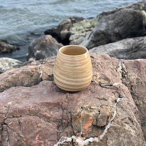 Op de afbeelding: Een kleine, handgemaakte keramische vaas met een lichtbruin en crèmekleurig gestreept patroon. De vaas is cilindrisch met een licht uitlopende rand. Hij staat op een roodachtig bruine rots met de oceaan op de achtergrond.