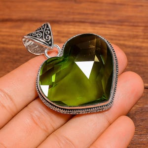 May include: A heart-shaped pendant with a large, faceted, olive-green gemstone. The gemstone is set in a silver-colored metal frame with intricate detailing. The pendant has a decorative bail for a chain.