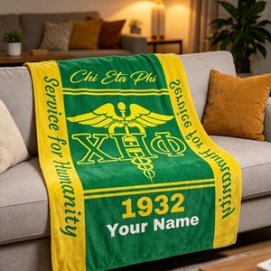 Op de afbeelding: Een groene en gele deken met de tekst "Chi Eta Phi" en "Service for Humanity". De deken heeft een wapen met vleugels en een caduceus, samen met het jaar "1932" en ruimte voor een naam. De deken is over een grijze bank gedrapeerd.