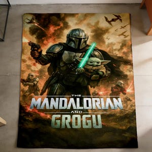 May include: A rectangular blanket featuring a scene from The Mandalorian. The image shows the Mandalorian holding Grogu with a green lightsaber. The blanket has a fiery background with the words "The Mandalorian and Grogu".