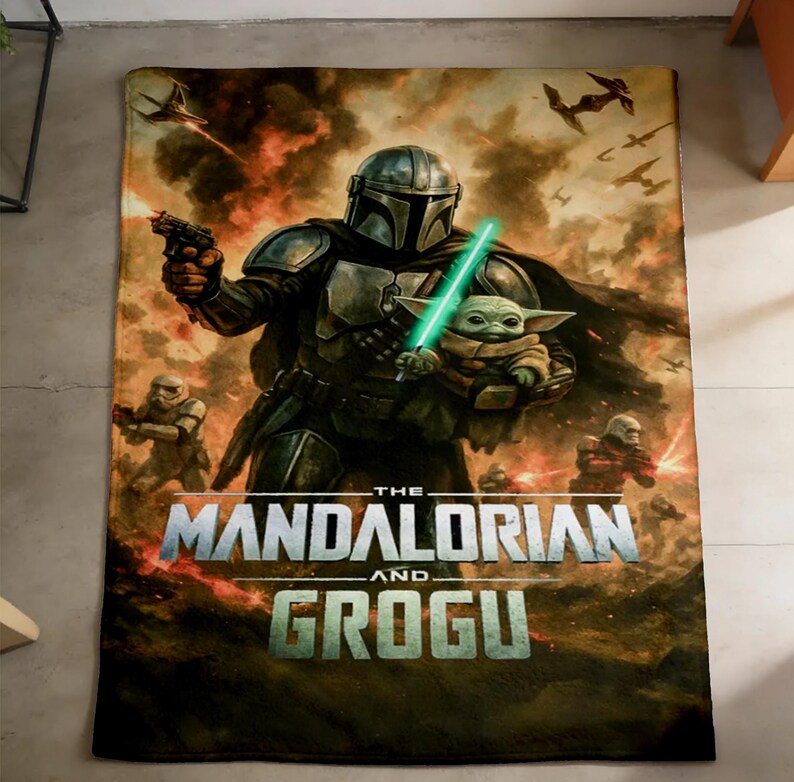 May include: A rectangular blanket featuring a scene from The Mandalorian. The image shows the Mandalorian holding Grogu with a green lightsaber. The blanket has a fiery background with the words "The Mandalorian and Grogu".