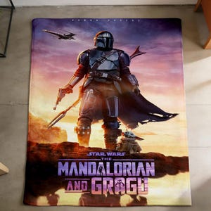 May include: A rectangular blanket featuring a scene from "The Mandalorian." The design showcases the Mandalorian in armor, a blaster, and Grogu. The blanket has a sunset background with a spaceship and the text "STAR WARS THE MANDALORIAN AND GROGU."