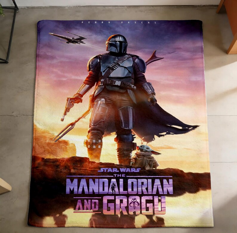 May include: A rectangular blanket featuring a scene from "The Mandalorian." The design showcases the Mandalorian in armor, a blaster, and Grogu. The blanket has a sunset background with a spaceship and the text "STAR WARS THE MANDALORIAN AND GROGU."