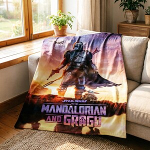 May include: A Star Wars themed blanket featuring The Mandalorian and Grogu. The blanket displays a detailed image of the Mandalorian in his armor, holding a weapon, with Grogu at his feet. The blanket has a sunset color scheme with the words "The Mandalorian and Grogu".