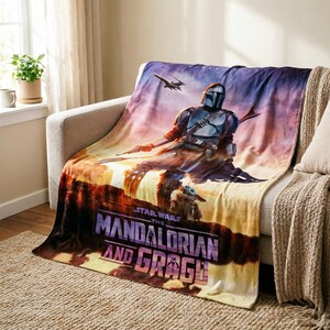 May include: A plush blanket featuring a scene from "The Mandalorian" with the Mandalorian and Grogu. The blanket has a sunset background with a spaceship. The words "The Mandalorian and Grogu" are printed on the bottom.