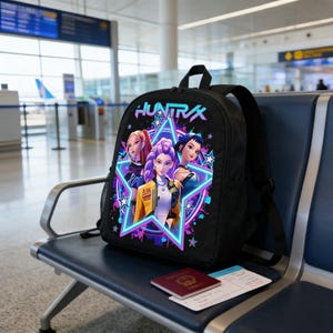 Może przedstawiać: Czarny plecak z kolorową grafiką przedstawiającą trzy kobiety w stylu anime wewnątrz neonowej gwiazdy, z napisem "HUNTRAX" powyżej. Plecak ma czarne paski i leży na niebieskiej ławce. Widoczny jest paszport i karta pokładowa.