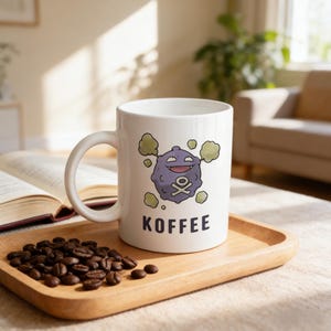 Può includere: Tazza in ceramica bianca con un disegno a cartoni animati di un personaggio viola con un teschio e ossa incrociate, circondato da nuvole verdi. La parola "KOFFEE" è stampata sotto. Chicchi di caffè su un vassoio di legno.