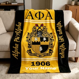 May include: A yellow and black blanket with the Greek letters ΑΦΑ in large black letters. The blanket features a crest with a helmet, crossed torches, and the text "1906 Your Name". The words "Alpha Phi Alpha" are written vertically.