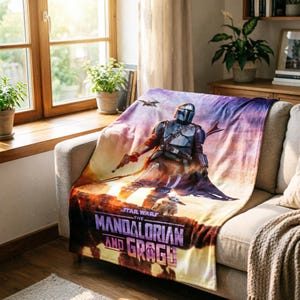 May include: A plush blanket featuring a vibrant graphic of The Mandalorian and Grogu from Star Wars. The design showcases the Mandalorian in armor, set against a sunset backdrop. The blanket is draped over a beige sofa.