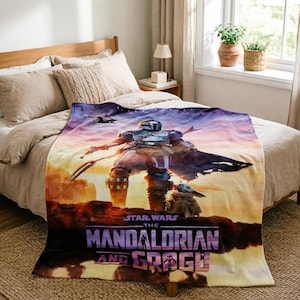 May include: A Star Wars The Mandalorian and Grogu blanket. The blanket features a colorful image of the Mandalorian and Grogu against a sunset backdrop. The blanket is draped over a bed with neutral-toned bedding.