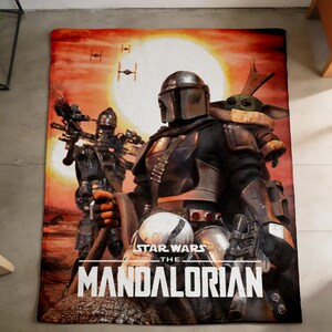 May include: A rectangular blanket featuring a scene from The Mandalorian. The image shows the Mandalorian, Grogu, and other characters against a sunset backdrop. The Star Wars and The Mandalorian logos are visible.