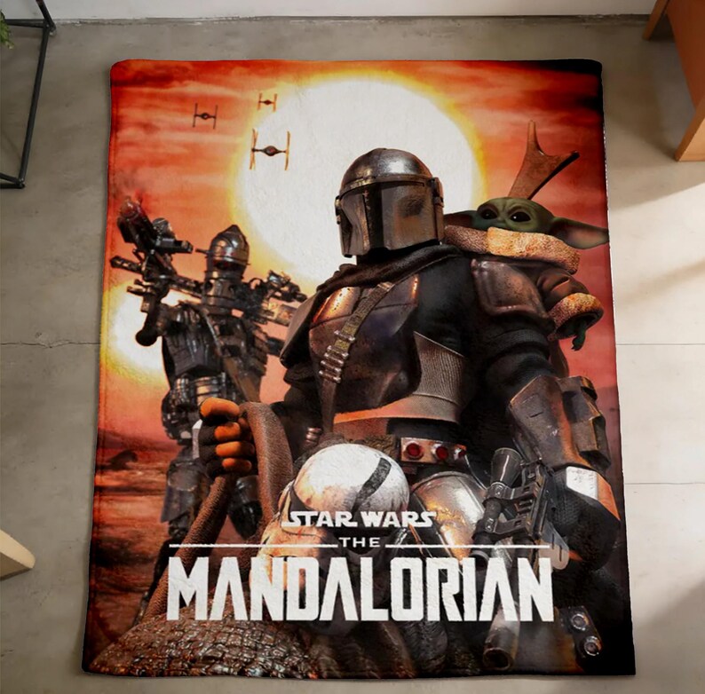May include: A rectangular blanket featuring a scene from The Mandalorian. The image shows the Mandalorian, Grogu, and other characters against a sunset backdrop. The Star Wars and The Mandalorian logos are visible.