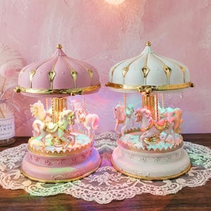 May include: Two carousel music boxes, one in pastel pink and one in white, each featuring gold-toned accents, a central pillar, and miniature horses suspended from the canopy. The bases are decorated with scalloped edges and gold trim.