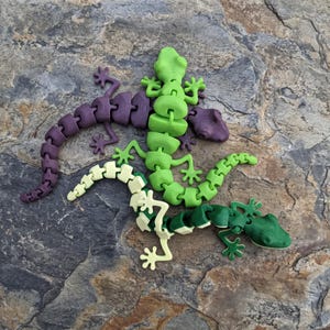 May include: Three articulated lizard figurines featuring segmented bodies that allow for flexible movement. One lizard is dark purple, one is bright lime green, and the third is a two-toned combination of light cream and dark forest green.