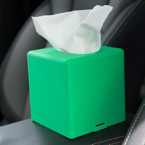 May include: A bright green tissue box with a square shape, featuring a slot for tissue dispensing. White tissues are visible, emerging from the top. The box is placed on a dark surface, possibly a car interior.