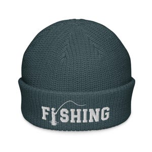 May include: A dark teal waffle-knit beanie with a folded cuff. The front features white embroidered text that reads FISHING, with the letter I replaced by a graphic of a person holding a fishing rod and line.
