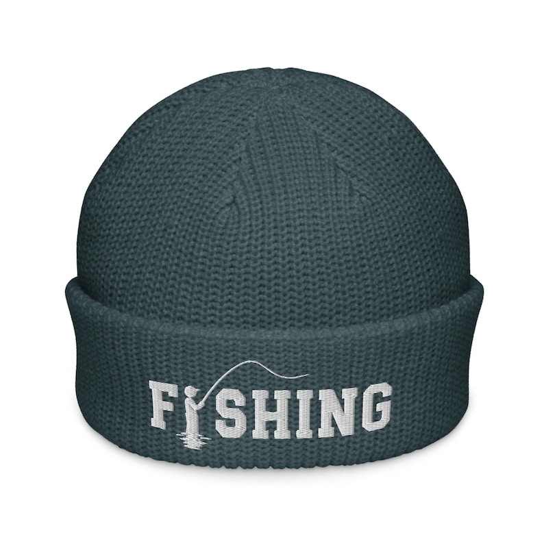 May include: A dark teal waffle-knit beanie with a folded cuff. The front features white embroidered text that reads FISHING, with the letter I replaced by a graphic of a person holding a fishing rod and line.