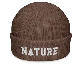May include: A brown knit beanie with a folded cuff. The word NATURE is embroidered in white block letters on the front of the cuff, with a small white pine tree icon replacing the letter A in the word.