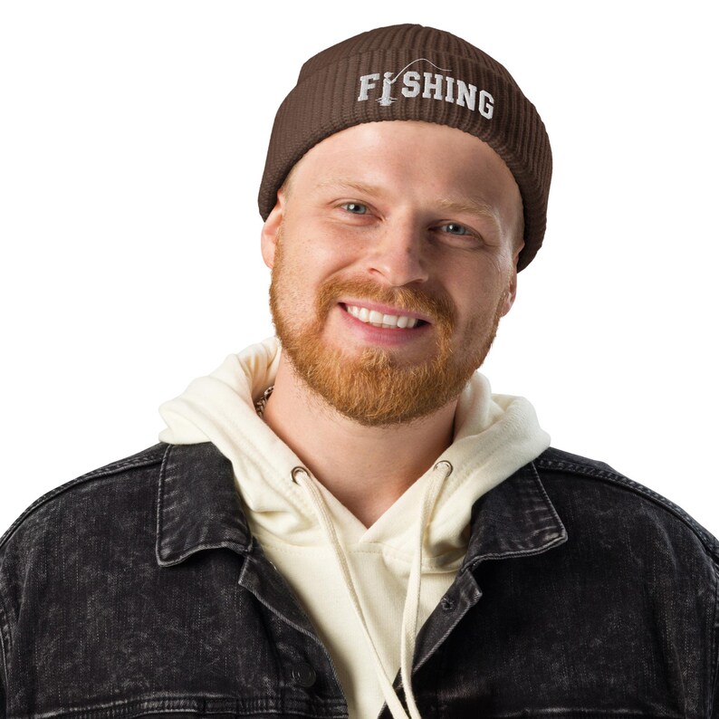 May include: A dark brown ribbed knit beanie featuring the white embroidered text FISHING, with the letter I replaced by a graphic of a fishing rod and line. The hat has a folded cuff and a textured, vertical knit pattern throughout the fabric.